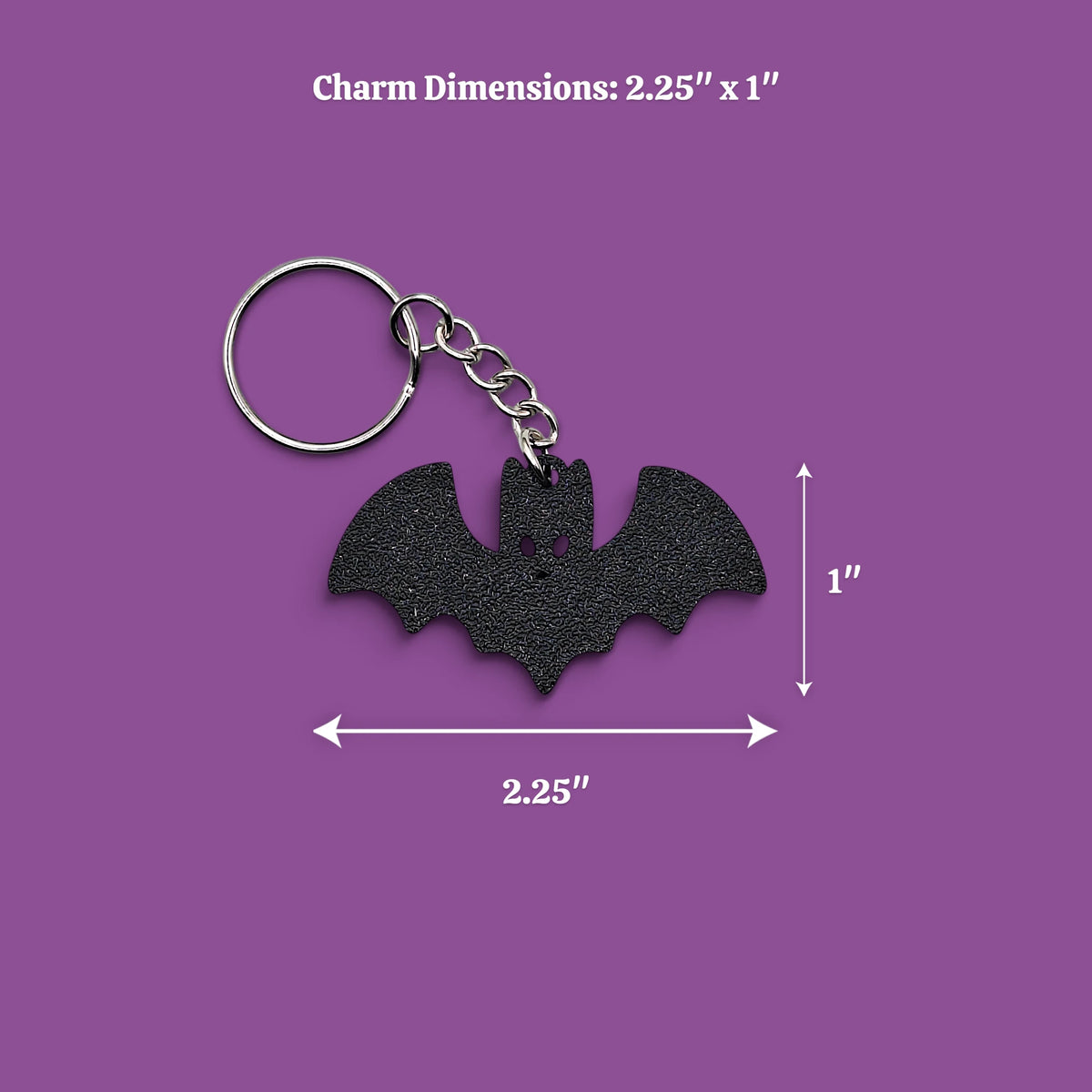 A black bat-shaped charm keychain with a silver metal split ring and chain, shown on a purple background with dimension arrows and text indicating 2.25 inches by 1 inch.