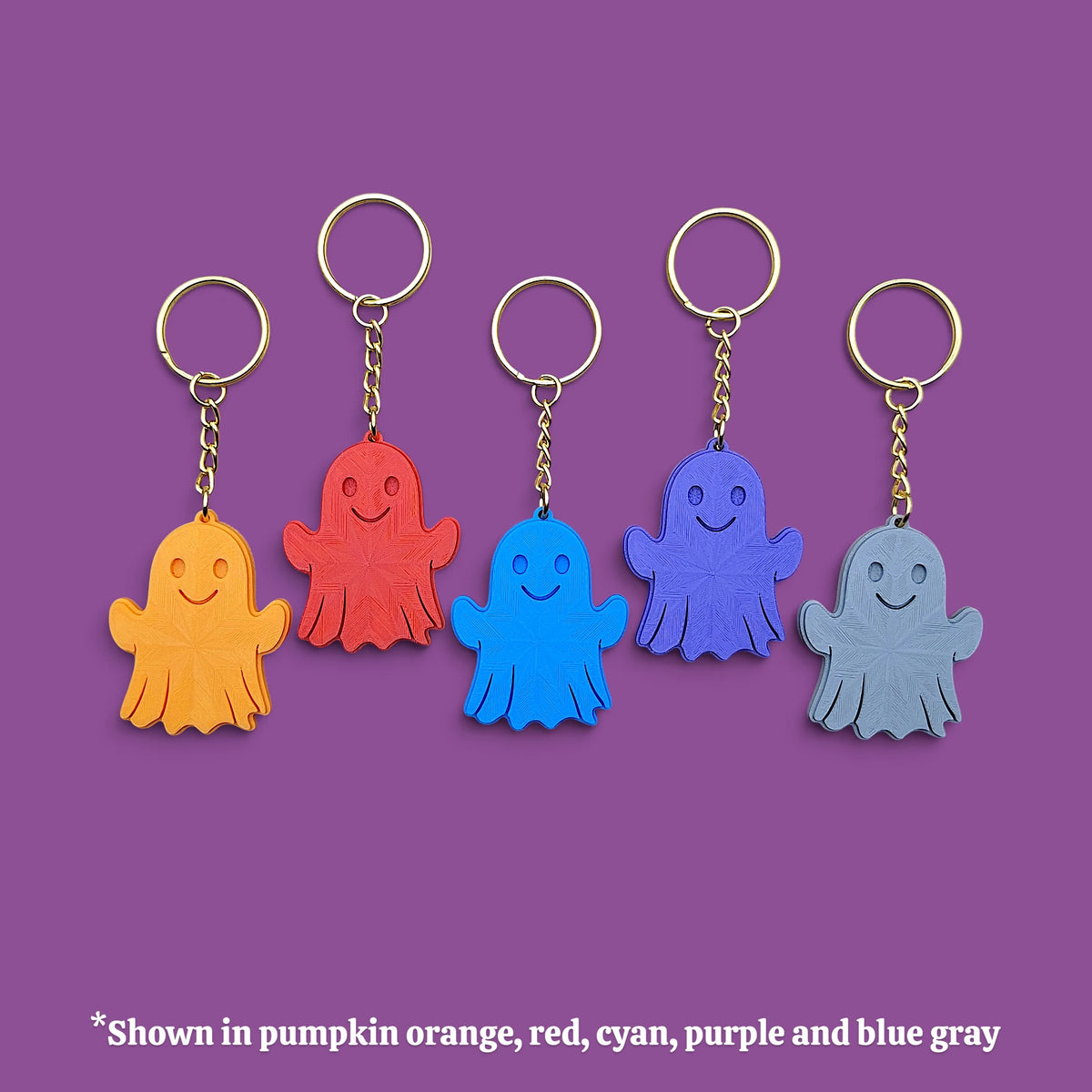 Five ghost-shaped keychains in different colors (orange, red, cyan, purple, and blue-gray) arranged in a row against a solid purple background, with a text note at the bottom indicating the color options.