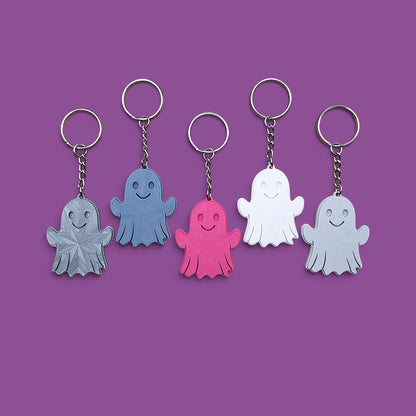 Five ghost-shaped keychains in different colors (gray, blue, pink, white, and silver) arranged in a row against a solid purple background.