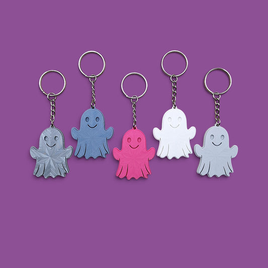 Five ghost-shaped keychains in different colors (gray, blue, pink, white, and silver) arranged in a row against a solid purple background.