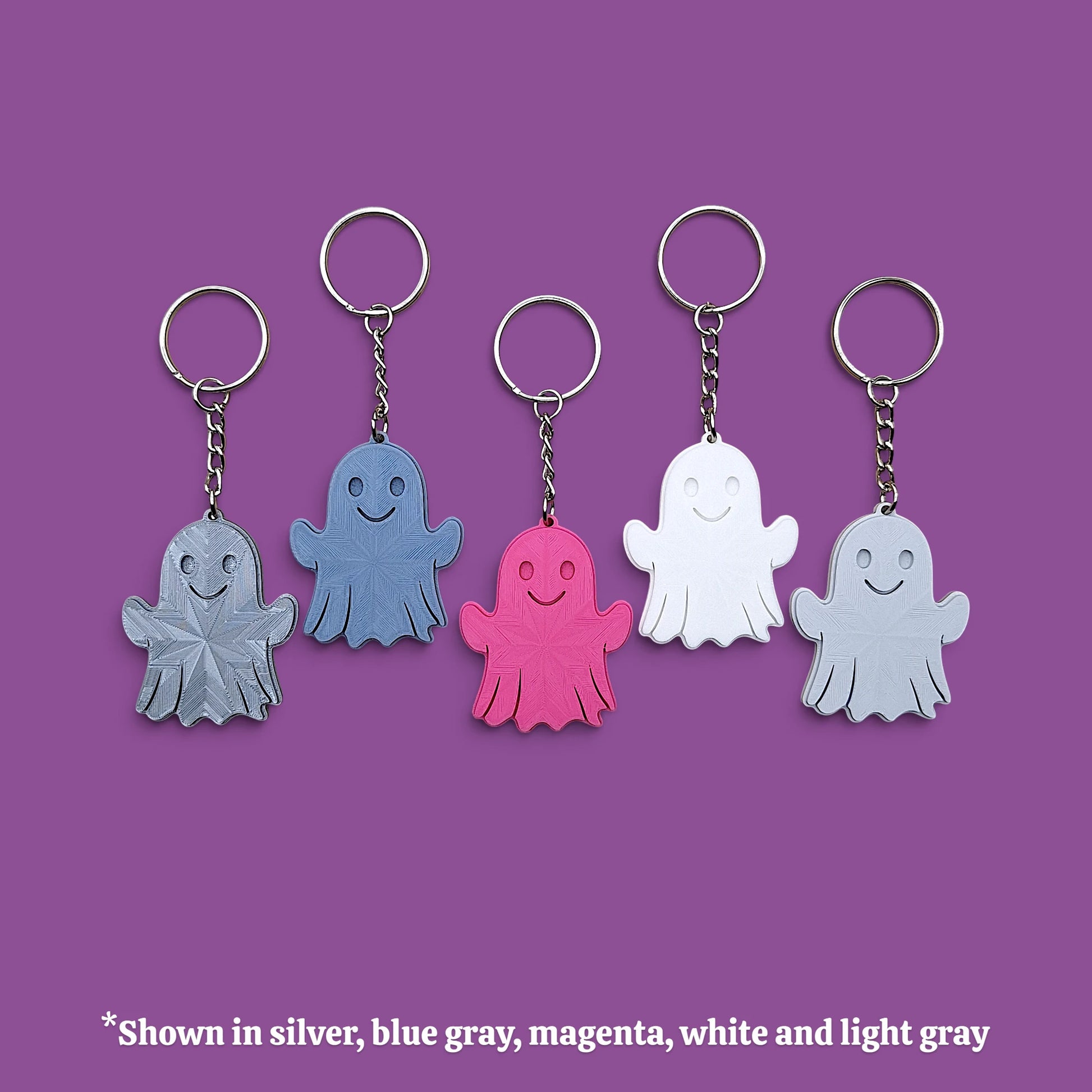 Five ghost-shaped keychains in different colors (silver, blue-gray, magenta, white, and light gray) arranged in a row against a solid purple background, with a text overlay at the bottom explaining the color options.