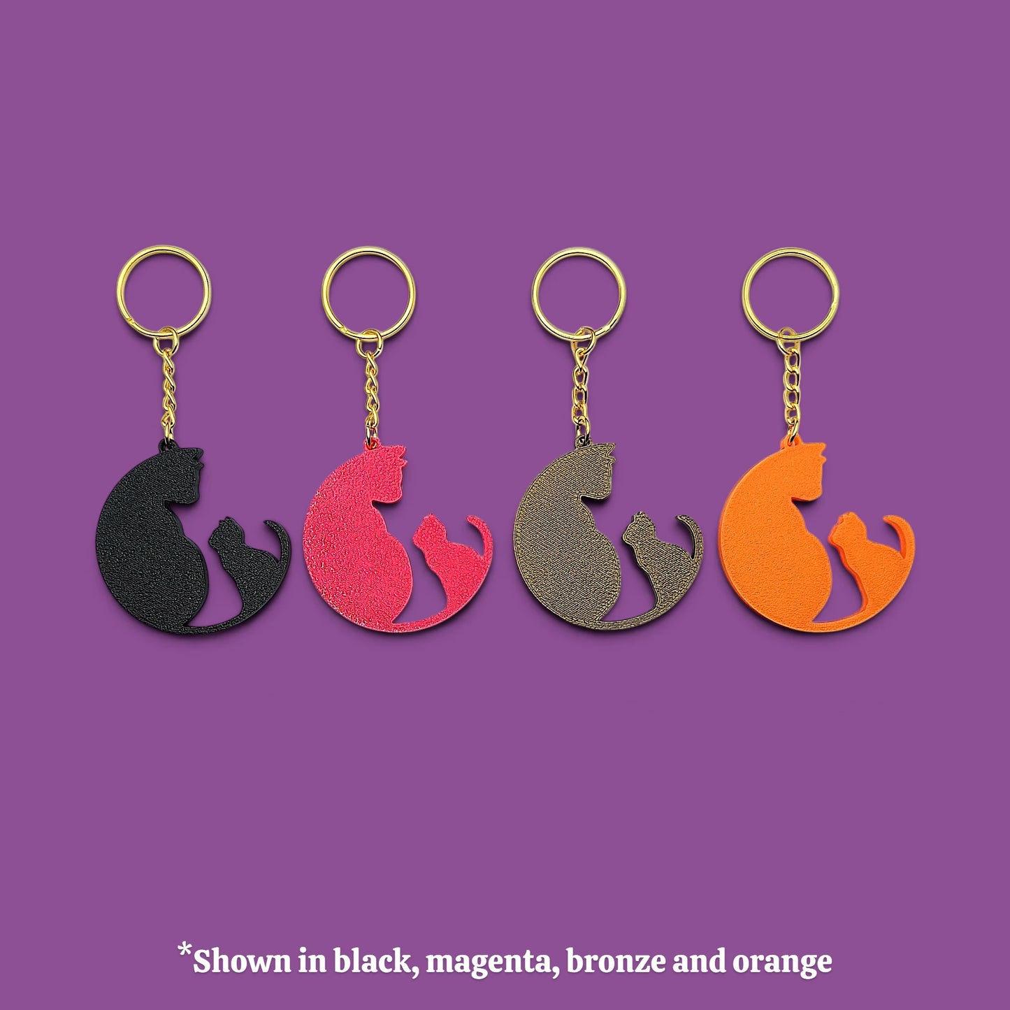 Four cat-shaped keychains in different colors (black, magenta, bronze, and orange) arranged in a row on a solid purple background, with a text overlay at the bottom stating '*Shown in black, magenta, bronze and orange'.