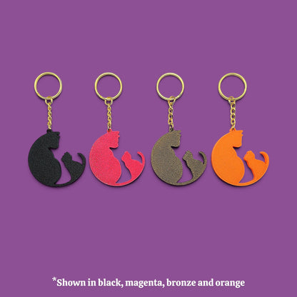 Four cat-shaped keychains in different colors (black, magenta, bronze, and orange) arranged in a row on a solid purple background, with a text overlay at the bottom stating '*Shown in black, magenta, bronze and orange'.