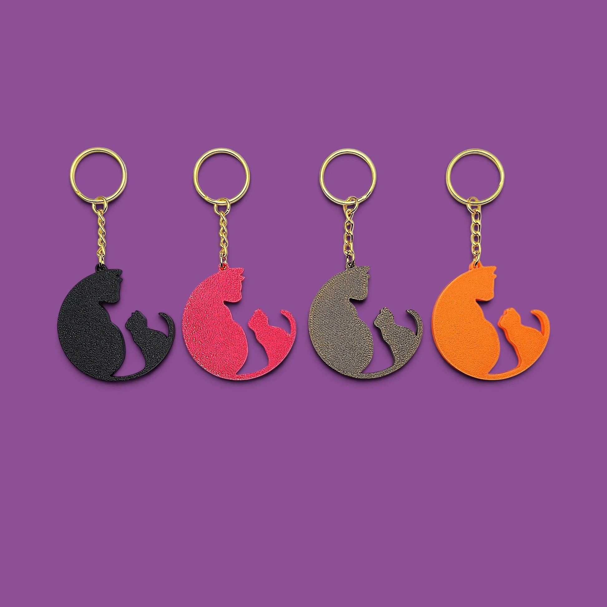 
Four cat-shaped keychains in different colors (black, magenta, bronze, and orange) arranged in a row on a solid purple background.
