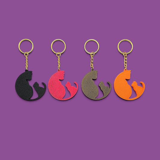 
Four cat-shaped keychains in different colors (black, magenta, bronze, and orange) arranged in a row on a solid purple background.