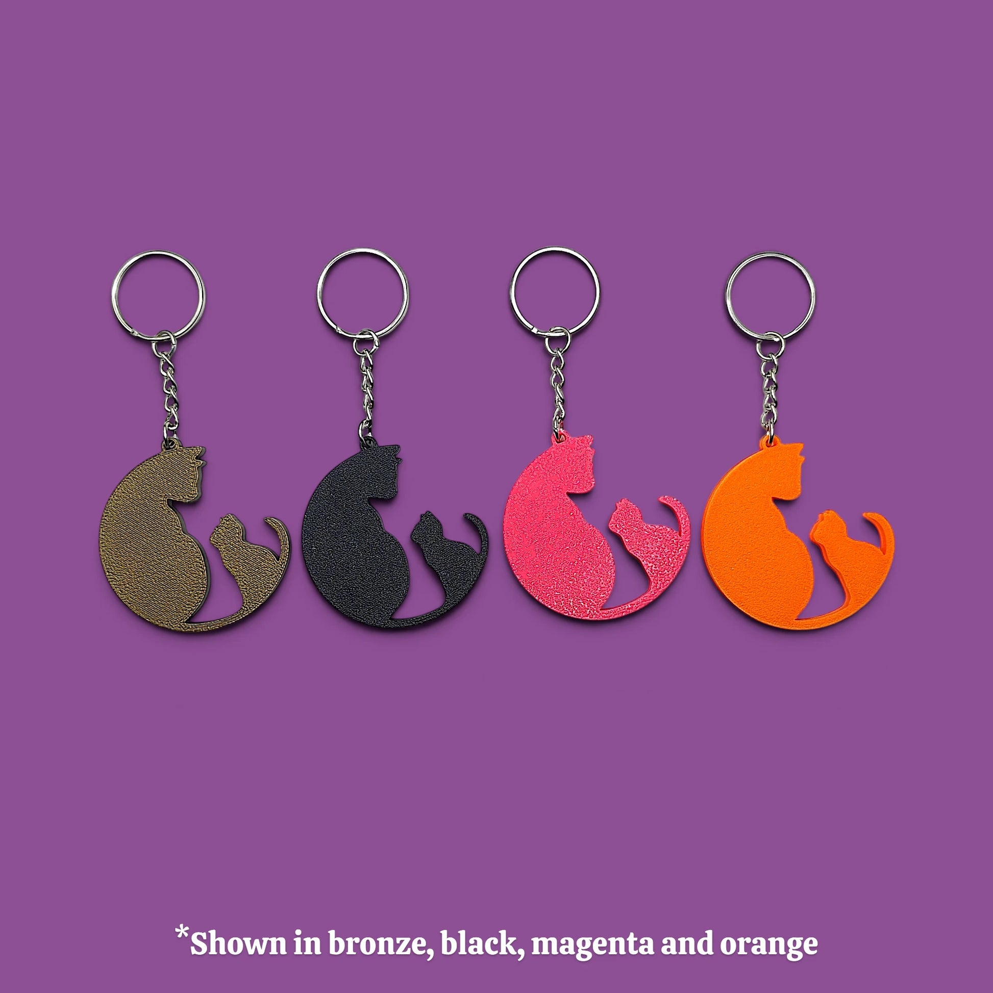 Four metal keychains shaped like a mother cat and her kitten in different colors (bronze, black, magenta, and orange) arranged in a row against a solid purple background, with a text overlay at the bottom stating '*Shown in bronze, black, magenta and orange'.