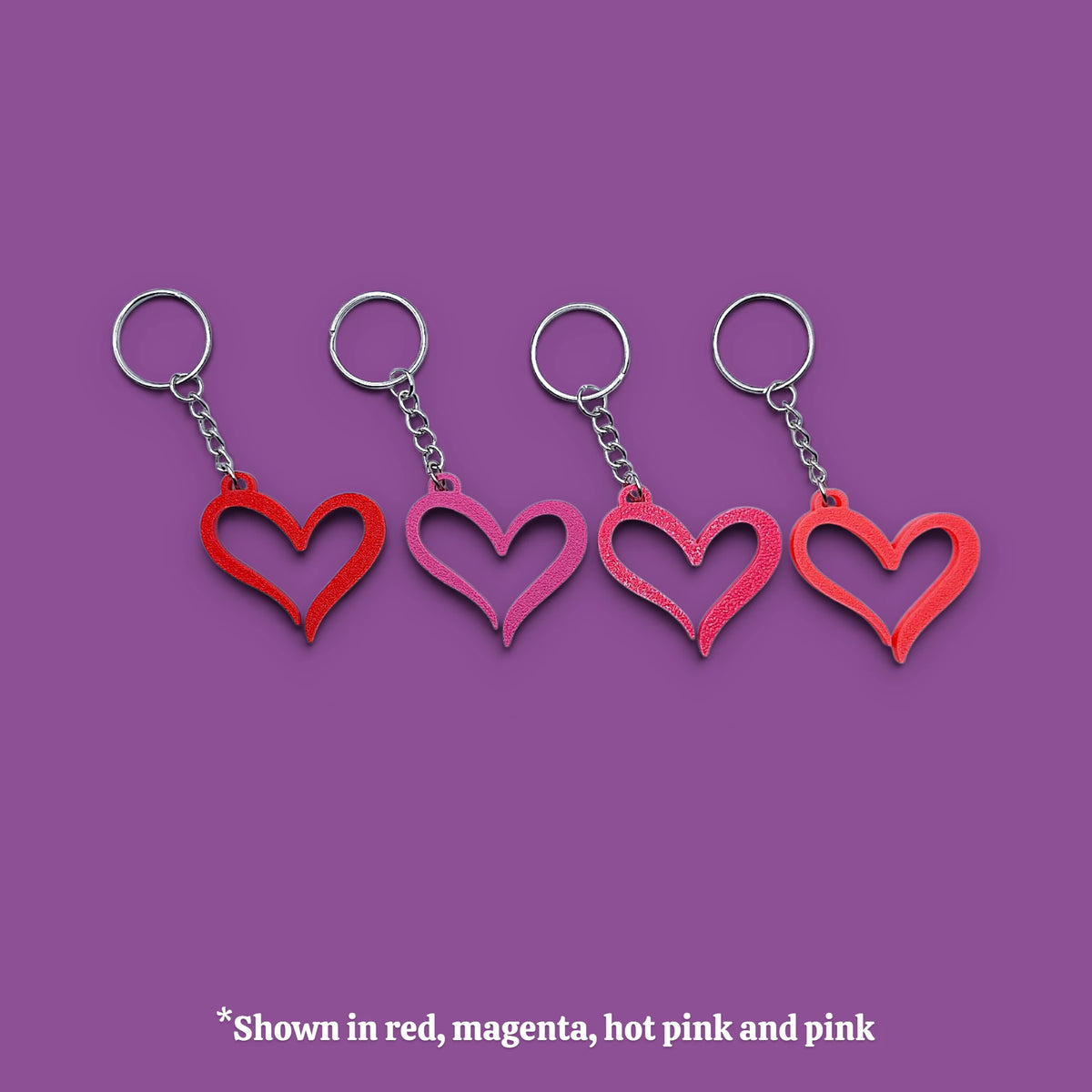 Four heart-shaped keychains in red, magenta, hot pink, and pink, arranged in a row on a solid purple background with a text note at the bottom.