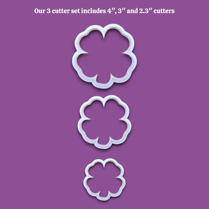 Three white four leaf clover shamrock  cookie cutters in sizes 4 inches, 3 inches, and 2.3 inches, displayed vertically on a solid gray background with black text at the top reading 'Our 3 cutter set includes 4", 3" and 2.3" cutters'