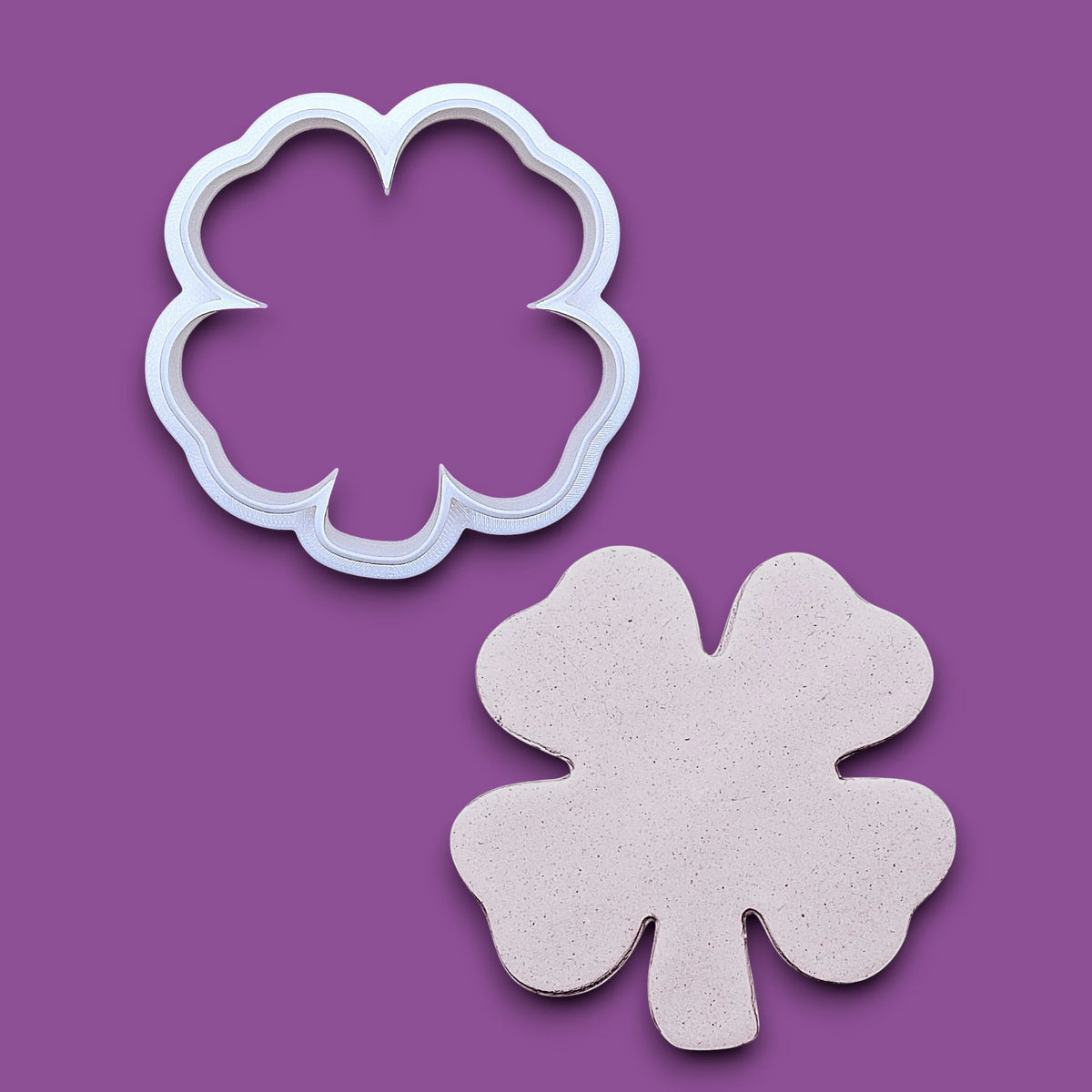 A white plastic four-leaf clover cookie cutter shown above its matching cutout on a solid gray background.