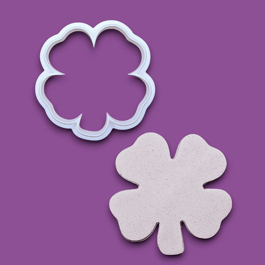 A white plastic four-leaf clover cookie cutter shown above its matching cutout on a solid gray background.