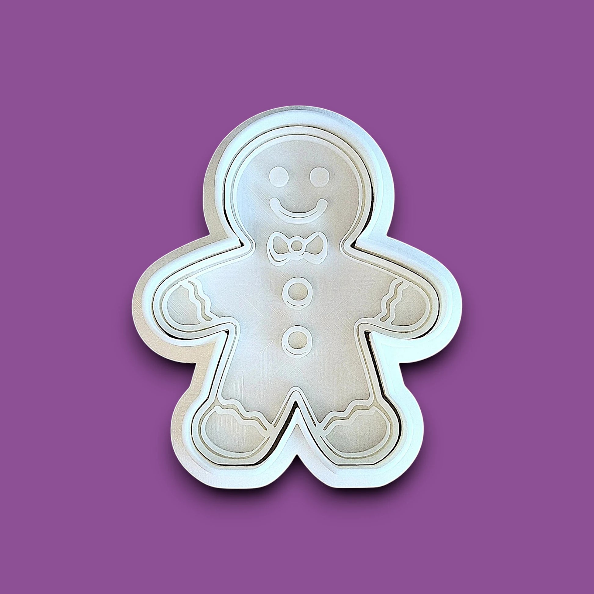 A white plastic gingerbread man cookie cutter with a smiling face and small bow tie detail, shown on a purple background.