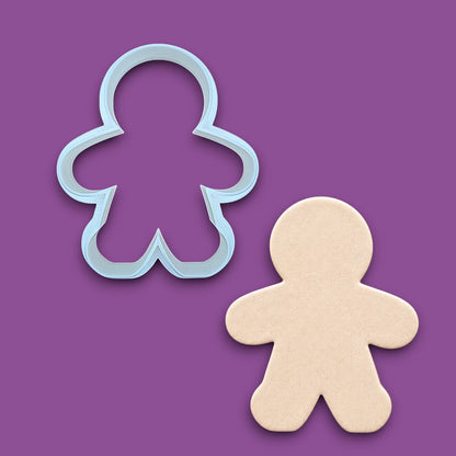 A white gingerbread man cookie cutter shown above a matching cutout of a gingerbread man made of speckled white cake mix, on a purple background.