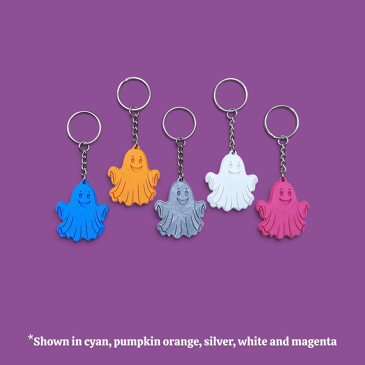 Five ghost-shaped keychains in different colors (cyan, pumpkin orange, silver, white, and magenta) arranged in a row on a solid purple background, with a text overlay at the bottom stating '*Shown in cyan, pumpkin orange, silver, white and magenta'.