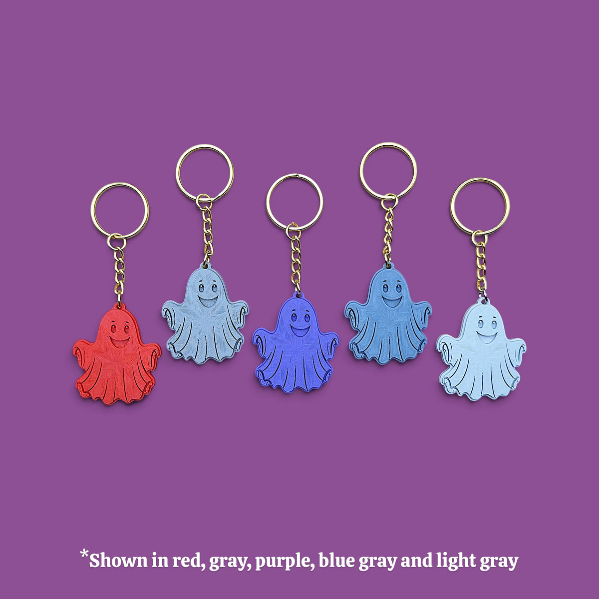 Five small ghost-shaped keychains with gold-tone rings and chains are arranged on a purple background. The ghosts come in red, gray, purple, blue-gray, and light gray colors.