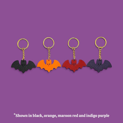 Four bat-shaped keychains in different colors (black, orange, maroon red, and indigo purple) lined up on a solid purple background with a text note at the bottom.