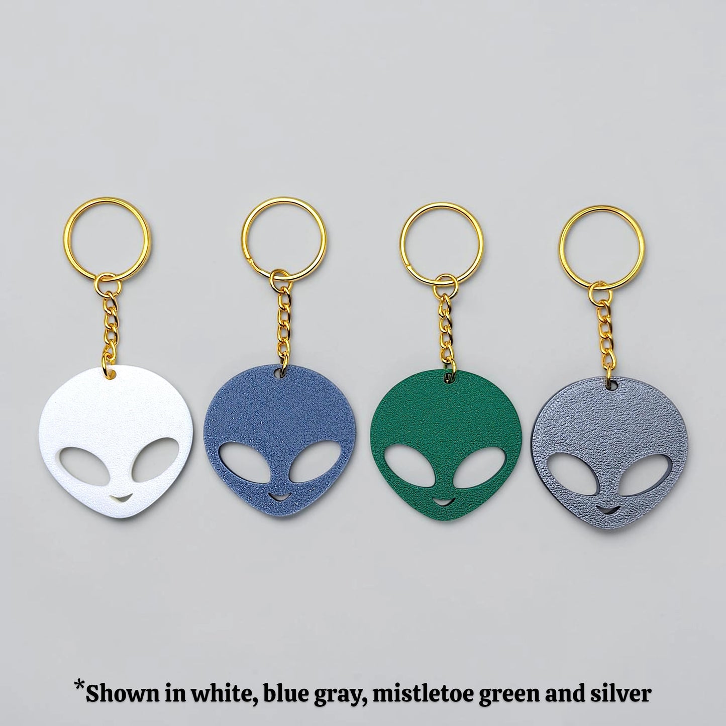 Four alien head keychains in white, blue gray, mistletoe green, and silver on a gray background.