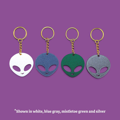 Four alien head keychains in different colors (white, blue-gray, green, and silver) with gold-tone rings and chains, displayed on a solid purple background with a text overlay at the bottom.