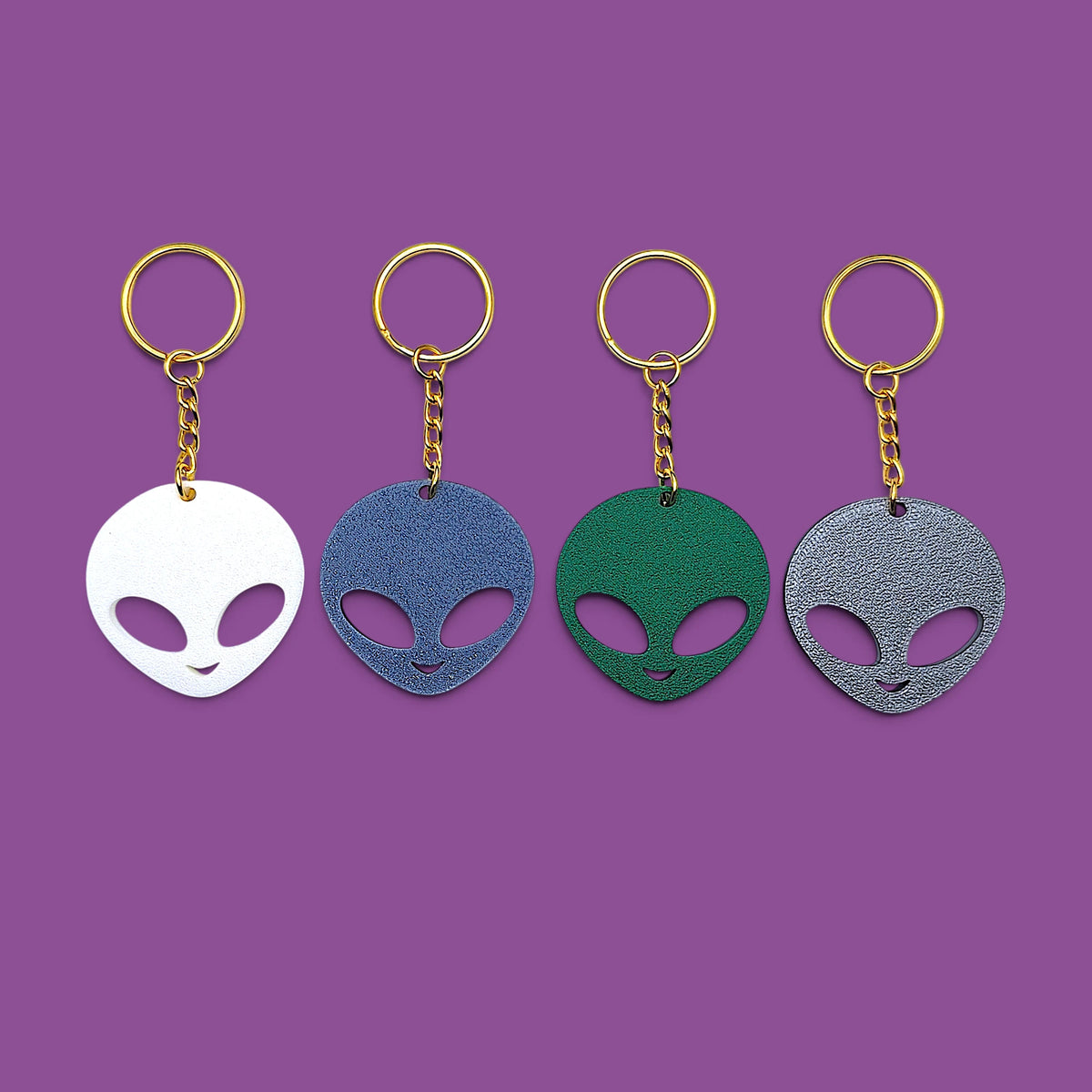 Four alien head keychains in white, blue, green, and gray, each with a gold-tone ring and chain, arranged in a row against a solid purple background.