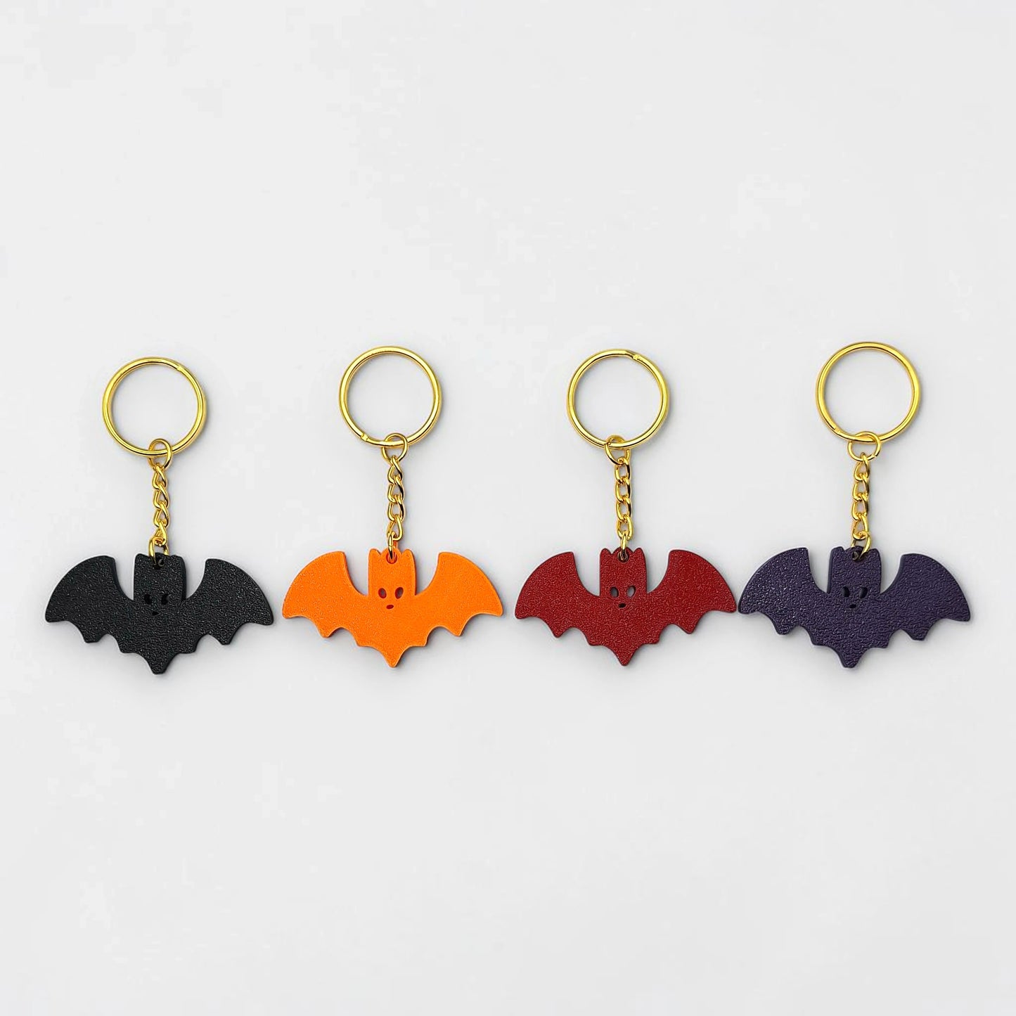 Four bat-shaped keychains in black, orange, red, and purple with gold chains on a white background.