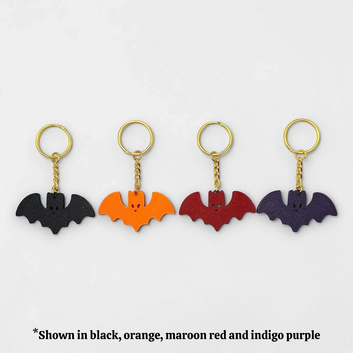 Four bat-shaped keychains in black, orange, maroon red, and indigo purple on a white background.