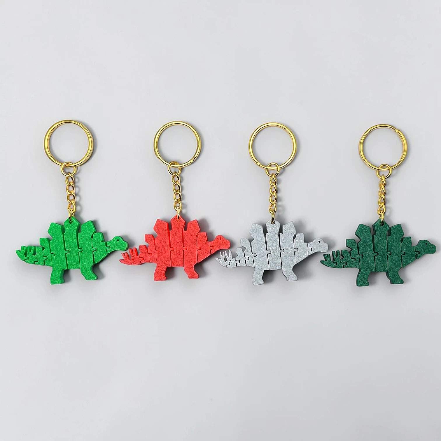 Four dinosaur-shaped keychains in green, red, gray, and green on a light gray background.