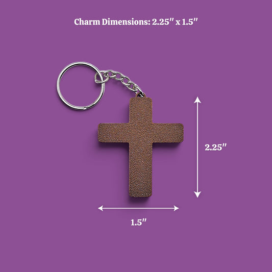 A small cross-shaped keychain charm in a textured brown finish with a silver metal key ring and chain, shown on a purple background with white dimension arrows and text reading 'Charm Dimensions: 2.25" x 1.5"'
