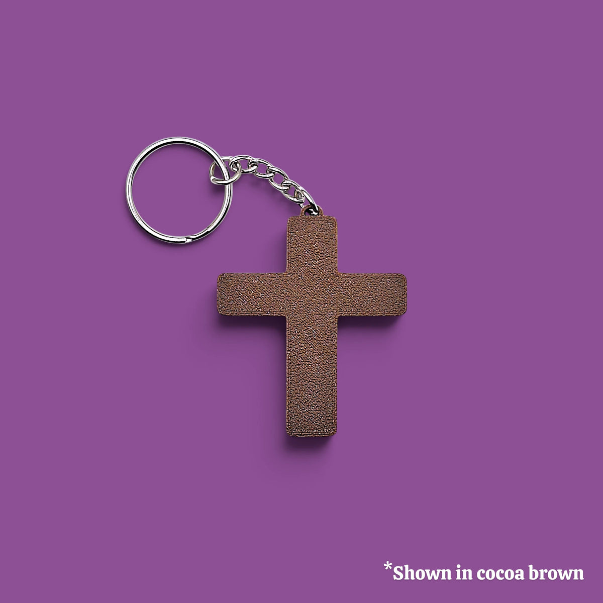 A textured cocoa brown cross-shaped keychain with a silver metal split ring and chain, displayed on a solid purple background with a small text note in the bottom right corner.