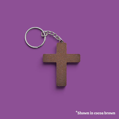A textured cocoa brown cross-shaped keychain with a silver metal split ring and chain, displayed on a solid purple background with a small text note in the bottom right corner.