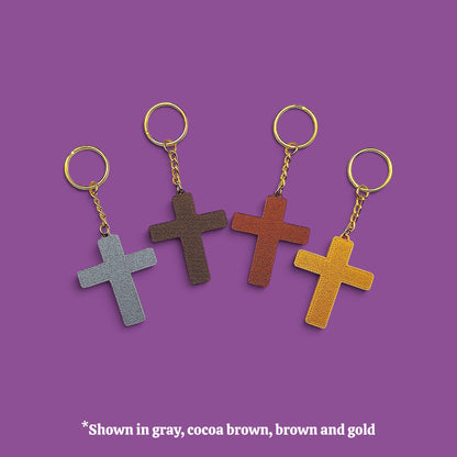 Four textured cross keychains in gray, cocoa brown, brown, and mustard yellow, each with gold-tone rings and chains, displayed on a solid purple background with a text overlay at the bottom.
