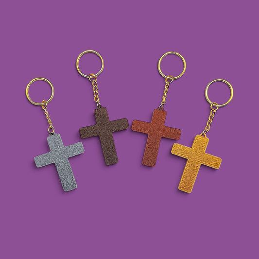 Four small cross keychains in different colors (gray, brown, reddish-brown, and mustard yellow) with gold-tone rings and chains, arranged on a solid purple background.