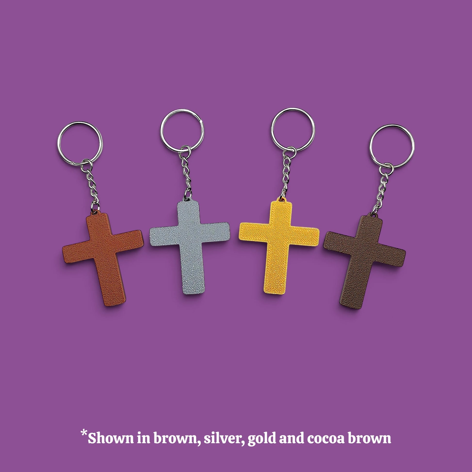 Four cross-shaped keychains in different colors (brown, silver, gold, and dark brown) arranged in a row on a solid purple background, with a text overlay at the bottom stating '*Shown in brown, silver, gold and cocoa brown'.