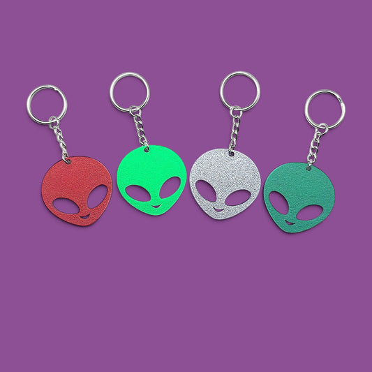 Four alien head keychains in red, green, silver, and teal, arranged in a row against a solid purple background.