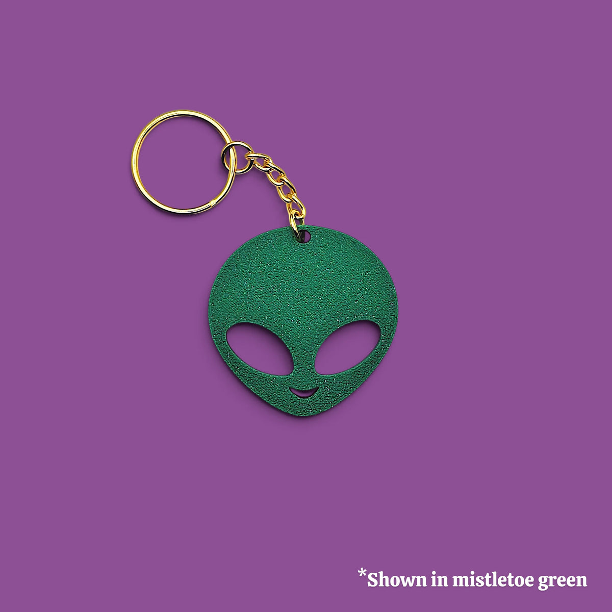 A green alien head keychain with a gold-tone split ring and chain, shown on a solid purple background with a small text note in the bottom right corner.