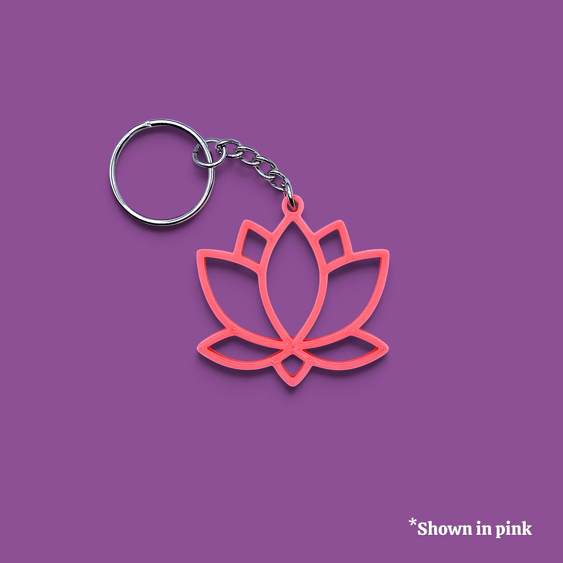 A pink lotus flower keychain with a silver metal split ring and chain, displayed on a solid purple background with a small text overlay in the bottom right corner that reads '*Shown in pink'.