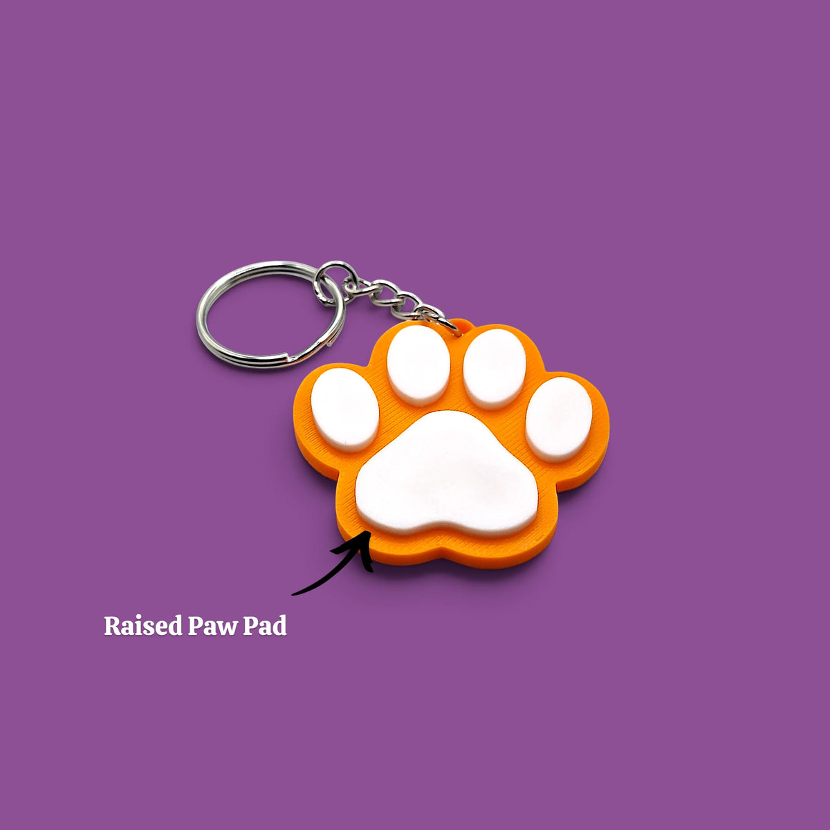An orange and white paw print keychain with a raised paw pad, shown on a solid purple background with a text label pointing to the pad.
