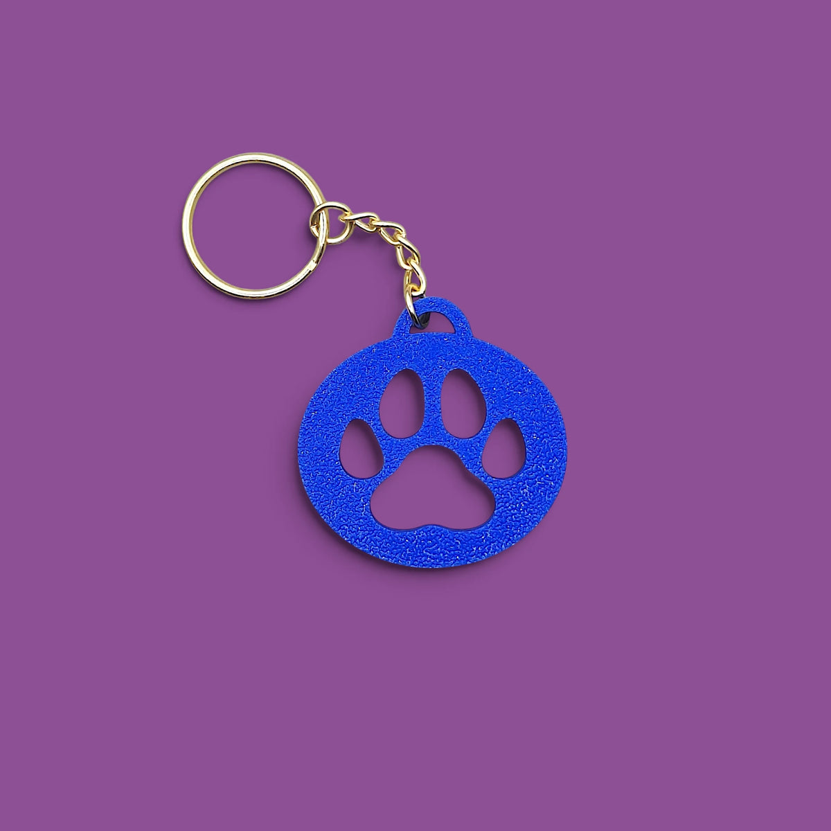 A blue paw print keychain with a gold metal split ring and chain, shown on a solid purple background.