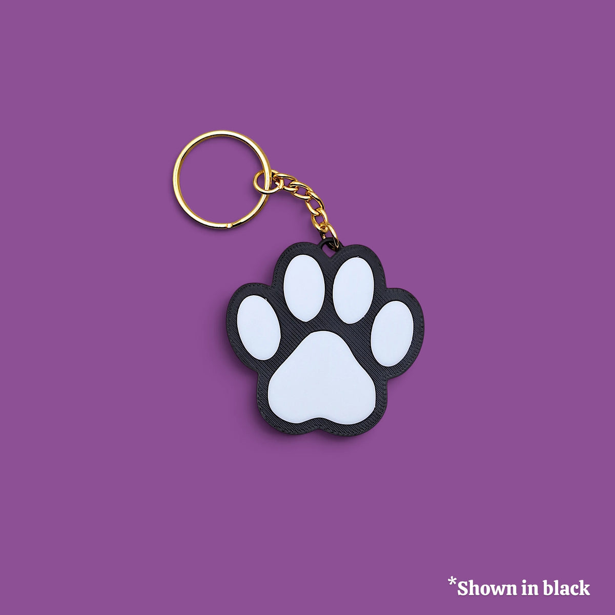 A paw print keychain with a black and white rubber paw pad and a gold split ring, shown on a solid purple background with a small text note in the bottom right corner that says '*Shown in black'.