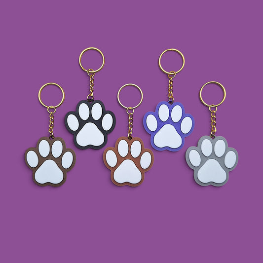 Five paw-shaped keychains in different colors (brown, black, blue, gray, and white) arranged in a row on a solid purple background.