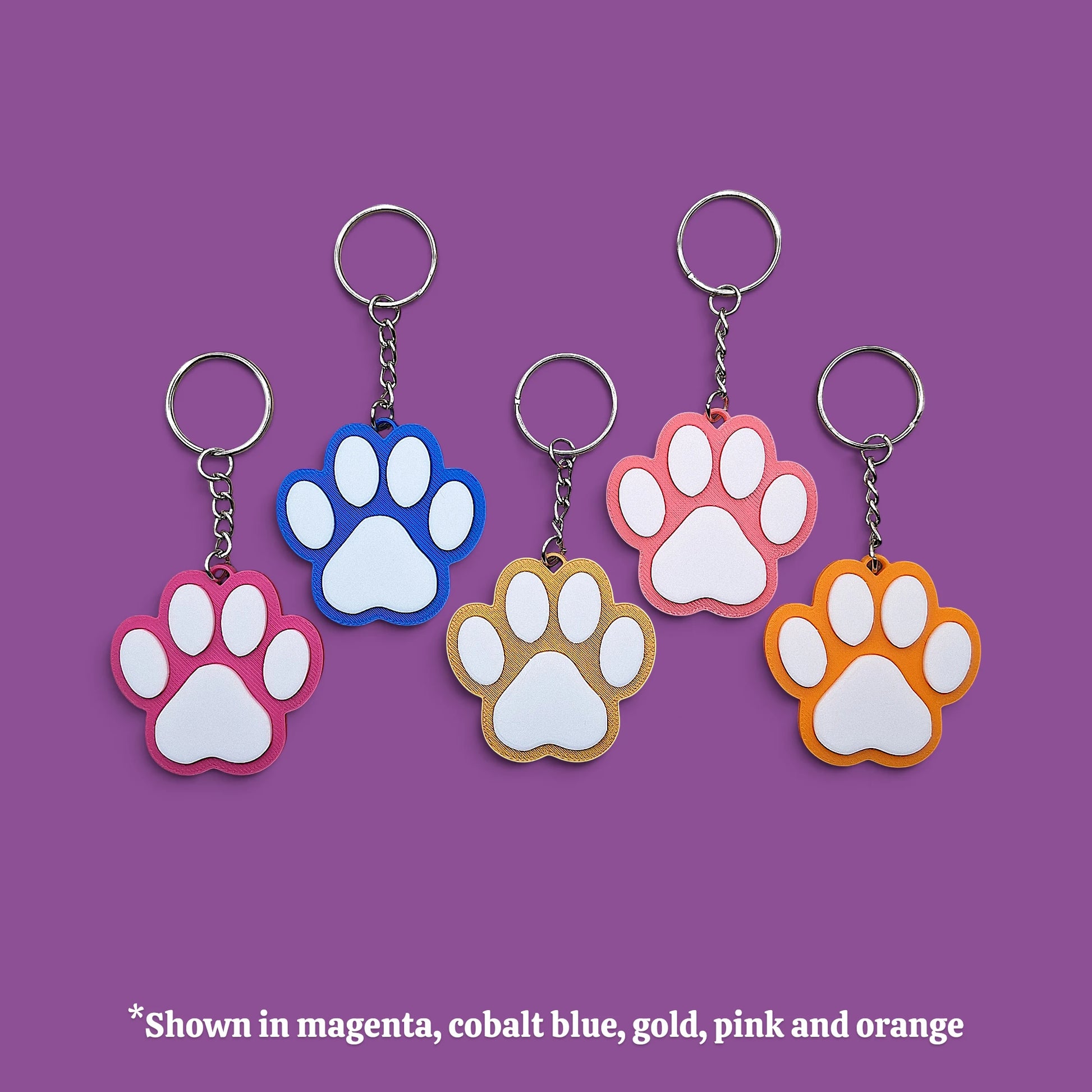 Five paw-shaped keychains in different colors (magenta, cobalt blue, gold, pink, and orange) arranged on a solid purple background, with a text overlay at the bottom stating '*Shown in magenta, cobalt blue, gold, pink and orange'.