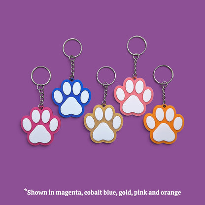 Five paw-shaped keychains in different colors (magenta, cobalt blue, gold, pink, and orange) arranged on a solid purple background, with a text overlay at the bottom stating '*Shown in magenta, cobalt blue, gold, pink and orange'.