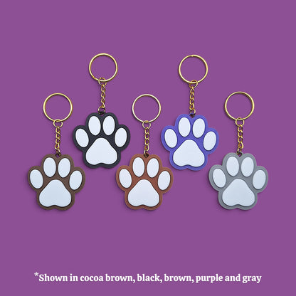 Five paw-shaped keychains with gold-tone rings and chains, shown in different colors (cocoa brown, black, brown, purple, and gray) against a solid purple background, with a text overlay at the bottom stating '*Shown in cocoa brown, black, brown, purple and gray'.