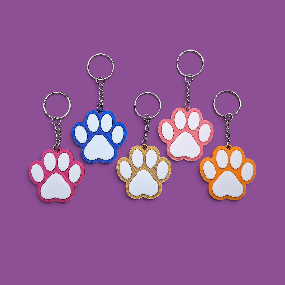 Five paw-print keychains in different colors (pink, blue, peach, tan, and orange) arranged on a solid purple background.
