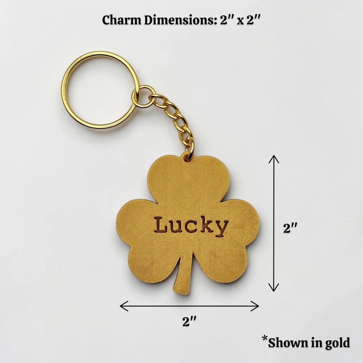 Gold shamrock keychain with 'Lucky' engraved in the center, shown with dimension lines indicating a 2x2 inch size on a light gray background.