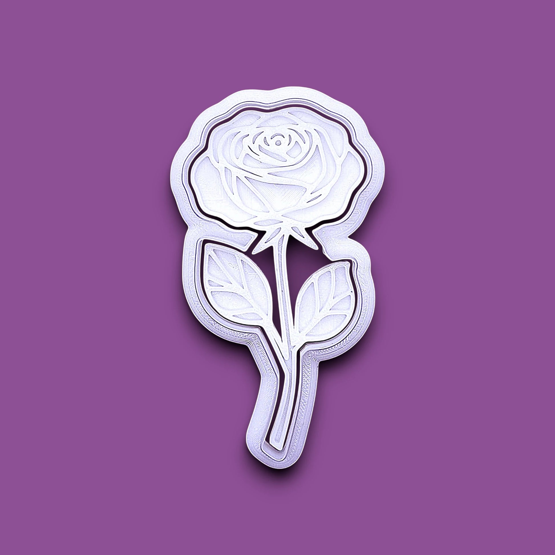 A white rose-shaped cookie cutter and stamp with layered petal and leaf details, shown on a solid purple background.