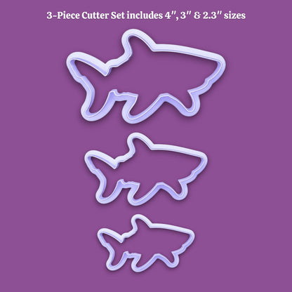 Three shark-shaped cookie cutters in different sizes displayed on a solid purple background, with white text at the top reading '3-Piece Cutter Set includes 4", 3", & 2.3" sizes'.