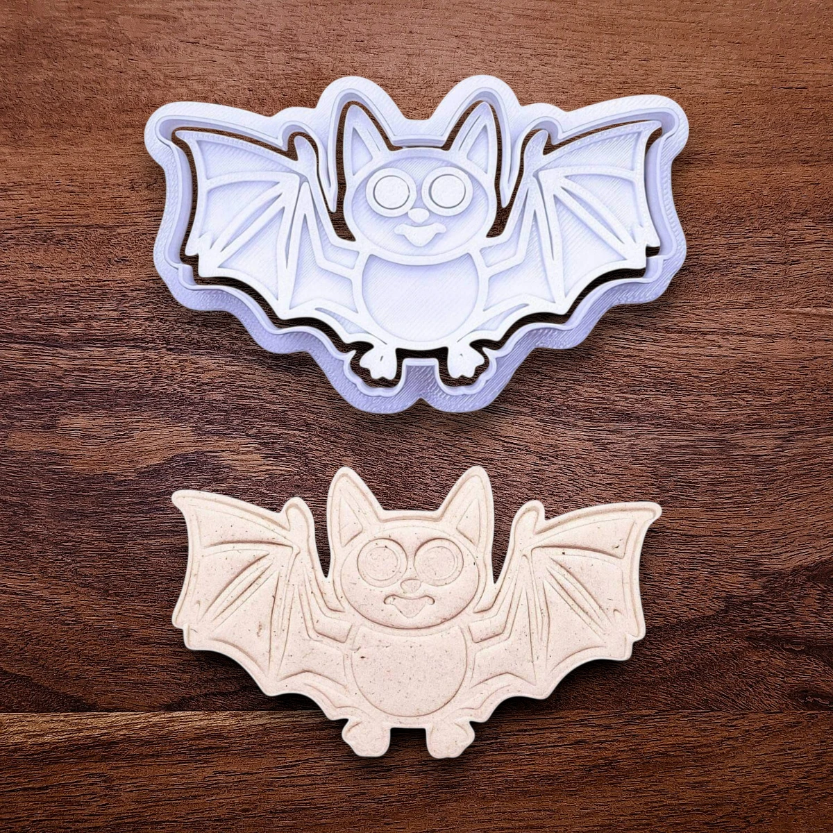 Bat cookie cutter with matching stamp and dough cutout with bat design pressed by cutter and stamp set, showing detailed lines—great for easy Halloween cookie decorating without icing.