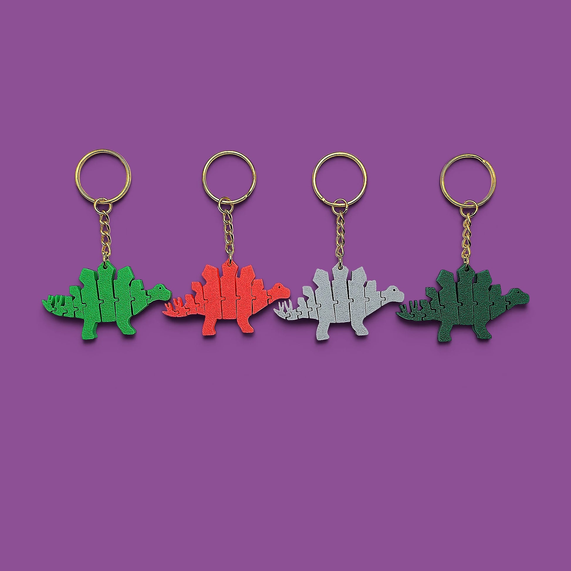 Four Stegosaurus keychains in green, red, silver, and dark green, arranged in a row on a solid purple background.