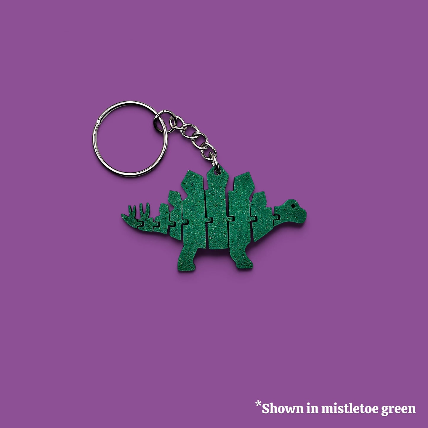 A green dinosaur-shaped keychain with a metal split ring and chain, shown on a solid purple background with a small text note in the bottom right corner.