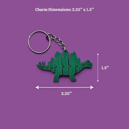 A green dinosaur-shaped keychain charm with a metal split ring and chain, shown on a purple background with dimension arrows and text reading Charm Dimensions: 2.25" x 1.5"