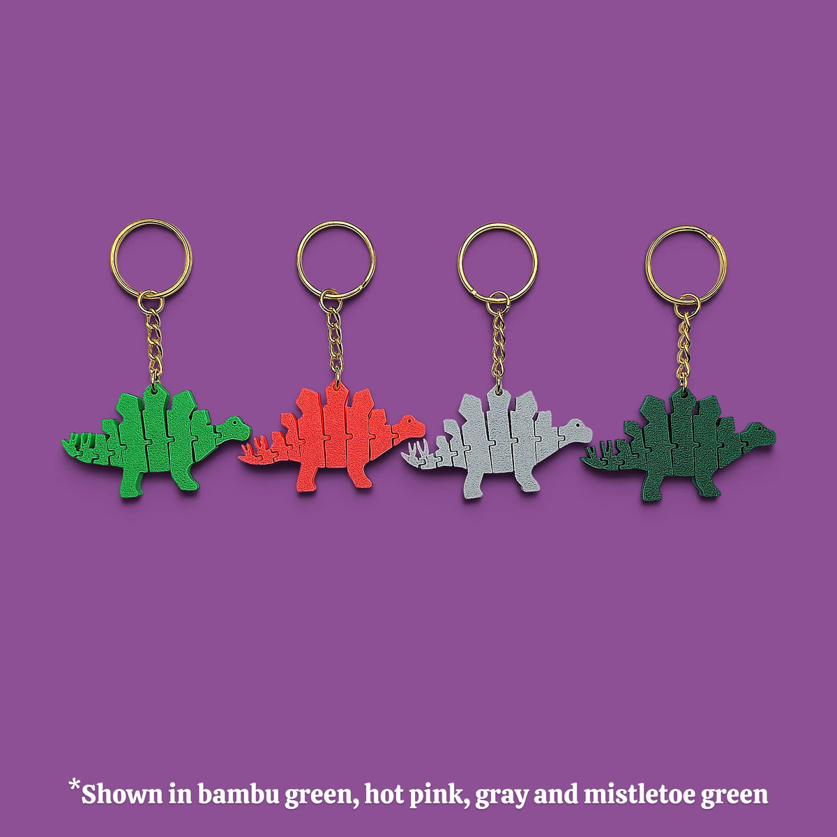 Four dinosaur-shaped keychains in different colors (bamboo green, hot pink, gray, and mistletoe green) lined up on a solid purple background with a text note at the bottom.